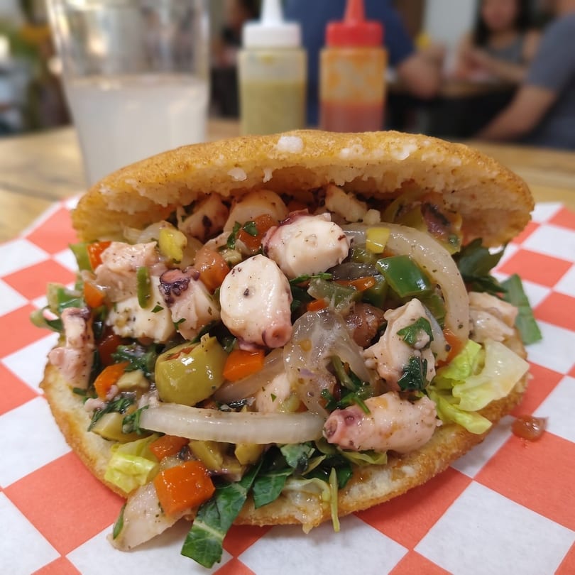 Close-up of a stuffed arepa with octopus, vegetables, and sauce on a red and white checkered paper.