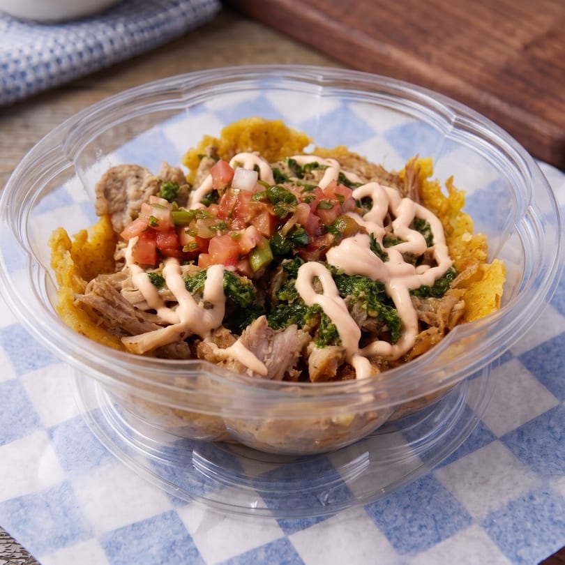 A clear plastic bowl filled with shredded meat, salsa, green sauce, and a creamy drizzle.